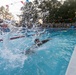 A swimmer in what appears to be military uniform or clothing is actively swimming in an outdoor competition pool, generating a large water splash in the foreground. A crowd of individuals dressed in military camouflage uniforms lines the poolside in the background, suggesting this is a military athletic event or competition. The scene is decorated with red, white, and blue pennant flags, indicating a formal or celebratory occasion.