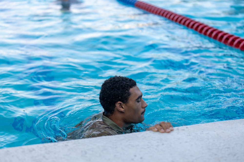 A person wearing camouflage military uniform is in a swimming pool, holding onto the pool edge. The setting appears to be a formal aquatic training facility with lane markers visible. This appears to be a military water survival or swim qualification exercise.