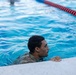 A person wearing camouflage military uniform is in a swimming pool, holding onto the pool edge. The setting appears to be a formal aquatic training facility with lane markers visible. This appears to be a military water survival or swim qualification exercise.