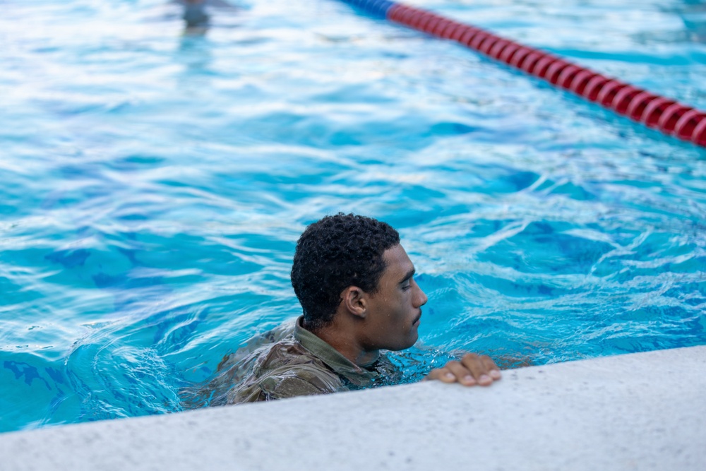 A person wearing military camouflage uniform is in a swimming pool, holding onto the pool edge. The individual appears to be participating in a military water training exercise, as evidenced by the fully clothed attire in the pool. The image is taken at close range, with lane dividers visible in the background.