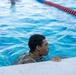 A person wearing military camouflage uniform is in a swimming pool, holding onto the pool edge. The individual appears to be participating in a military water training exercise, as evidenced by the fully clothed attire in the pool. The image is taken at close range, with lane dividers visible in the background.