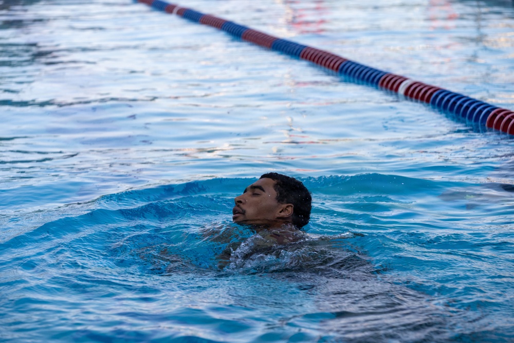 A young man is swimming in a competitive lap pool, with his head above water and eyes closed. The image captures him mid-stroke, surrounded by rippling blue water. A red and blue lane divider rope is visible in the background.