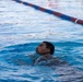 A young man is swimming in a competitive lap pool, with his head above water and eyes closed. The image captures him mid-stroke, surrounded by rippling blue water. A red and blue lane divider rope is visible in the background.