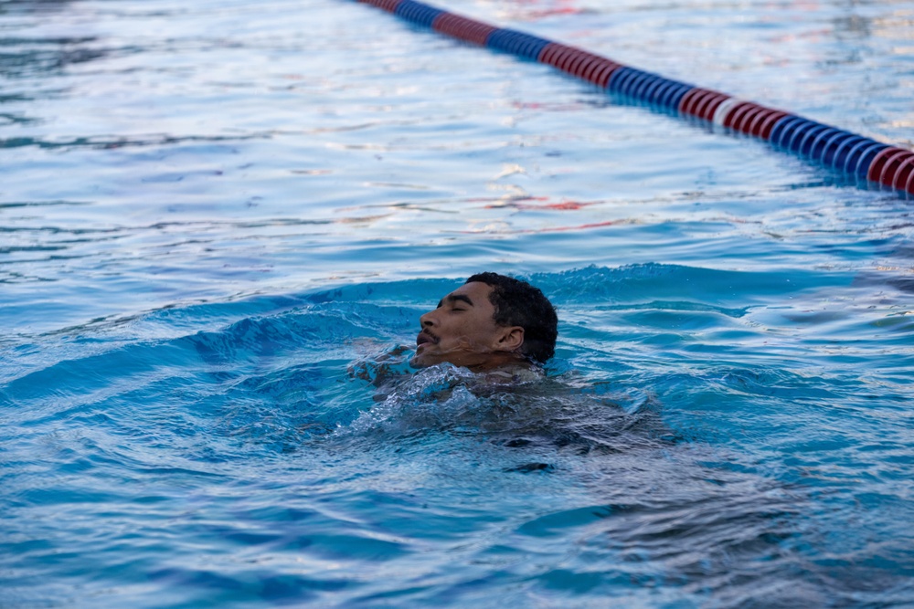 A person is swimming in a lap pool, with their head just above the water surface and eyes closed. The image captures the swimmer mid-stroke in what appears to be an outdoor or indoor competitive swimming facility. A red and blue lane divider is visible in the background.