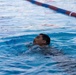 A person is swimming in a lap pool, with their head just above the water surface and eyes closed. The image captures the swimmer mid-stroke in what appears to be an outdoor or indoor competitive swimming facility. A red and blue lane divider is visible in the background.