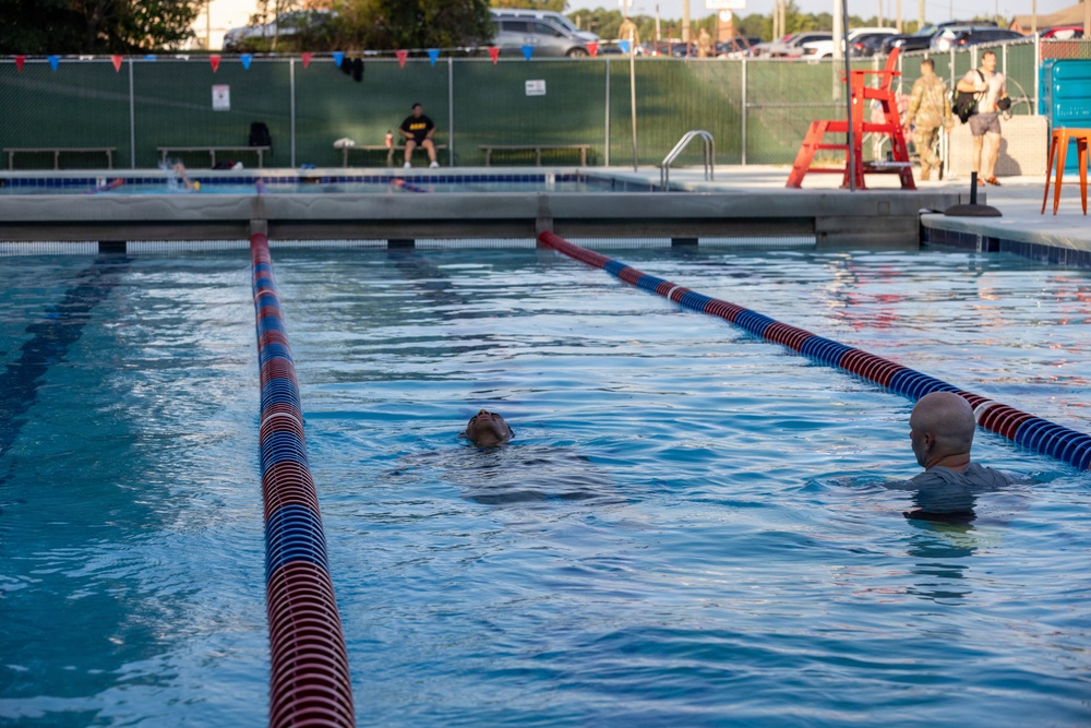 An outdoor swimming pool with lane dividers in red and blue is shown with two swimmers in the water. A lifeguard stand is visible in the background along with several observers on the pool deck. The setting appears to be a recreational or military facility pool based on the surrounding infrastructure.