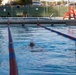 An outdoor swimming pool with lane dividers in red and blue is shown with two swimmers in the water. A lifeguard stand is visible in the background along with several observers on the pool deck. The setting appears to be a recreational or military facility pool based on the surrounding infrastructure.