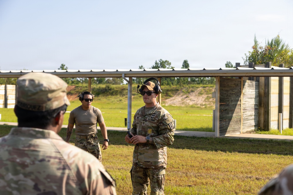 The image shows U.S. Army personnel at an outdoor shooting range facility. A soldier in full OCP (Operational Camouflage Pattern) uniform appears to be briefing or instructing a group, while another soldier in a tan t-shirt stands nearby. The setting includes a covered firing line structure with berms visible in the background.
