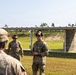 The image shows U.S. Army personnel at an outdoor shooting range facility. A soldier in full OCP (Operational Camouflage Pattern) uniform appears to be briefing or instructing a group, while another soldier in a tan t-shirt stands nearby. The setting includes a covered firing line structure with berms visible in the background.
