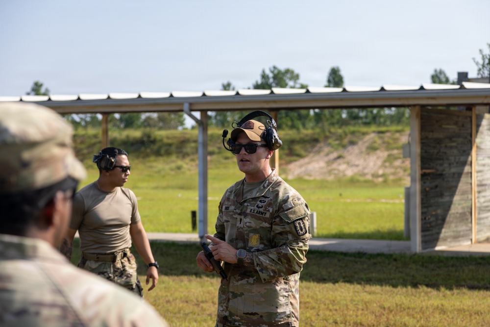 The image shows U.S. Army personnel at an outdoor shooting range, likely during a training exercise or qualification event. A soldier in full MultiCam uniform with 'U.S. ARMY' tape is speaking or instructing, wearing hearing protection and sunglasses. Additional personnel in military attire are visible in the background near a covered range structure.