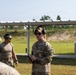 The image shows U.S. Army personnel at an outdoor shooting range, likely during a training exercise or qualification event. A soldier in full MultiCam uniform with 'U.S. ARMY' tape is speaking or instructing, wearing hearing protection and sunglasses. Additional personnel in military attire are visible in the background near a covered range structure.