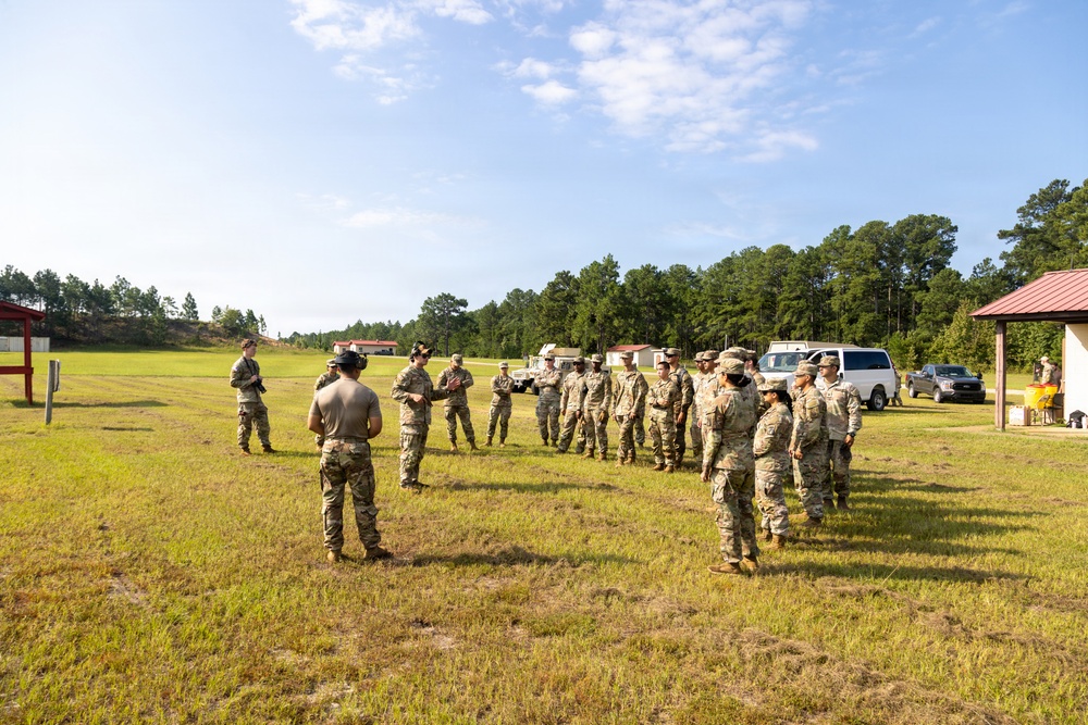 A group of military personnel in camouflage uniforms are assembled in an open grassy field, appearing to receive a briefing or instruction from a leader facing them. The setting appears to be a military training area or range facility, with vehicles, a covered structure, and tree lines visible in the background. The scene suggests a pre-training or after-action review formation.