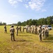 A group of military personnel in camouflage uniforms are assembled in an open grassy field, appearing to receive a briefing or instruction from a leader facing them. The setting appears to be a military training area or range facility, with vehicles, a covered structure, and tree lines visible in the background. The scene suggests a pre-training or after-action review formation.