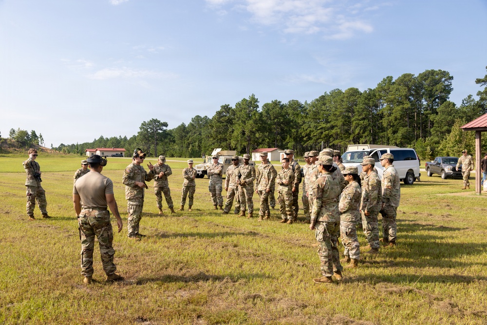 A group of military personnel in OCP (Operational Camouflage Pattern) uniforms are gathered in an open grassy field, appearing to receive a briefing or instruction from a leader. The setting appears to be a military training area or installation, with small buildings, a HMMWV (Humvee), and other vehicles visible in the background. The scene suggests a pre-mission briefing, training exercise, or range instruction.