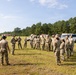 A group of military personnel in OCP (Operational Camouflage Pattern) uniforms are gathered in an open grassy field, appearing to receive a briefing or instruction from a leader. The setting appears to be a military training area or installation, with small buildings, a HMMWV (Humvee), and other vehicles visible in the background. The scene suggests a pre-mission briefing, training exercise, or range instruction.