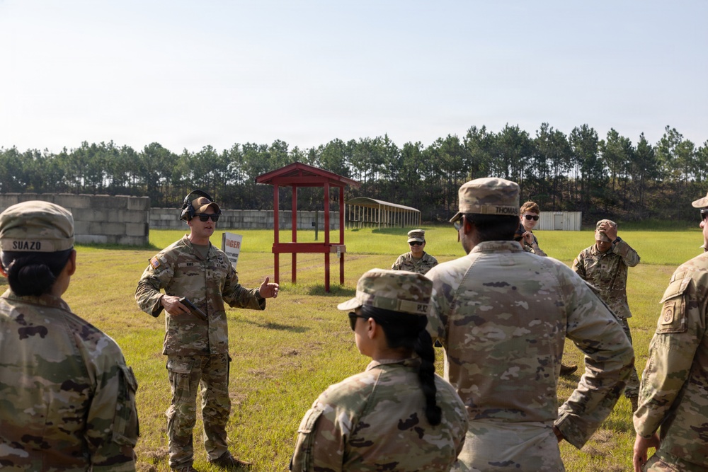 A group of military personnel in U.S. Army OCP (Operational Camouflage Pattern) uniforms are gathered at an outdoor firing range. One soldier appears to be conducting a briefing or safety instruction to the assembled group. The setting includes a red-roofed range control structure, concrete barriers, and a tree line in the background.