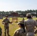 A group of military personnel in U.S. Army OCP (Operational Camouflage Pattern) uniforms are gathered at an outdoor firing range. One soldier appears to be conducting a briefing or safety instruction to the assembled group. The setting includes a red-roofed range control structure, concrete barriers, and a tree line in the background.