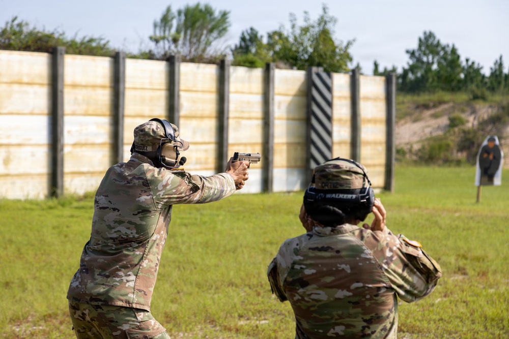 Two individuals in military camouflage uniforms are engaged in a pistol shooting exercise at an outdoor firing range. The shooter on the left is in an extended firing stance aiming a pistol downrange at silhouette targets, while the second individual appears to be observing or scoring. The range features a wooden barrier wall and standard silhouette targets in the background.