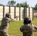 Two individuals in military camouflage uniforms are engaged in a pistol shooting exercise at an outdoor firing range. The shooter on the left is in an extended firing stance aiming a pistol downrange at silhouette targets, while the second individual appears to be observing or scoring. The range features a wooden barrier wall and standard silhouette targets in the background.