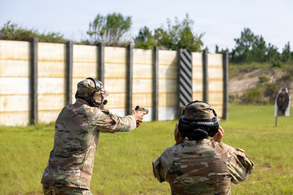 Two military personnel in OCP (Operational Camouflage Pattern) uniforms are at an outdoor shooting range. One soldier is actively firing a pistol downrange toward silhouette targets while the other observes or coaches from behind. Both are wearing hearing protection and eye protection consistent with range safety protocols.