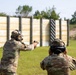 Two military personnel in OCP (Operational Camouflage Pattern) uniforms are at an outdoor shooting range. One soldier is actively firing a pistol downrange toward silhouette targets while the other observes or coaches from behind. Both are wearing hearing protection and eye protection consistent with range safety protocols.