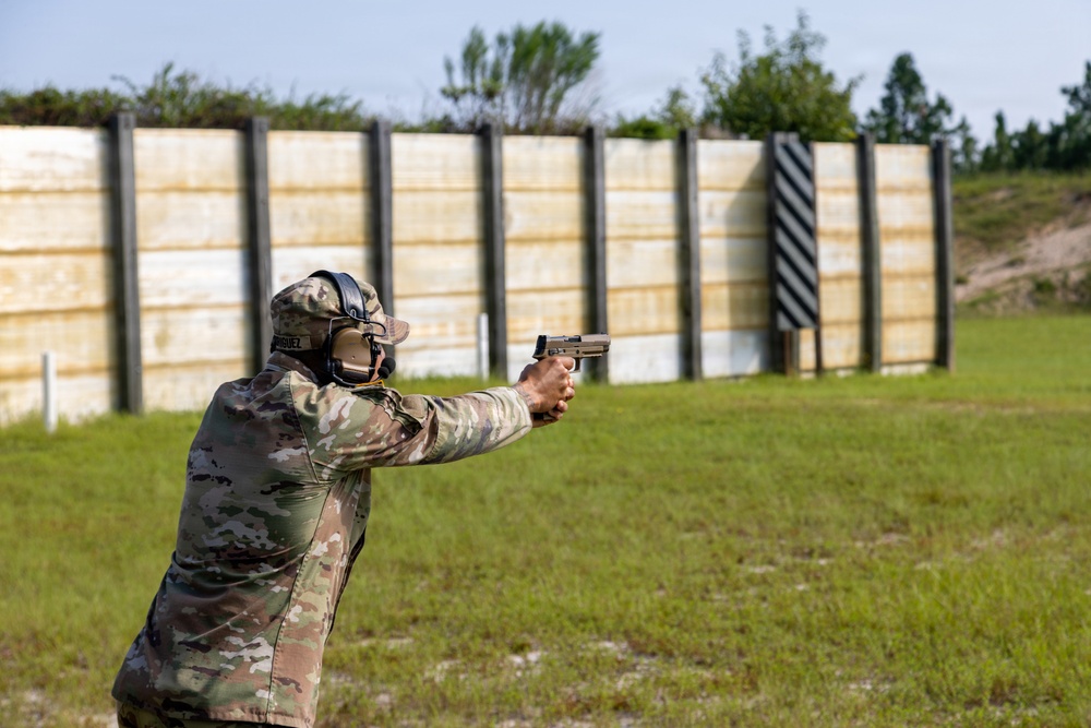 A person in military camouflage uniform is firing a pistol at an outdoor shooting range. The shooter is using a two-handed grip stance with arms extended toward a series of wooden target backer panels downrange. The setting appears to be a formal military or law enforcement training facility.