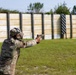 A person in military camouflage uniform is firing a pistol at an outdoor shooting range. The shooter is using a two-handed grip stance with arms extended toward a series of wooden target backer panels downrange. The setting appears to be a formal military or law enforcement training facility.