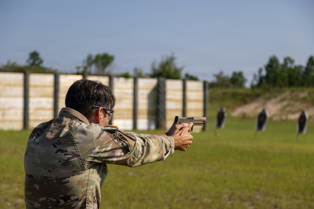 A person in military camouflage uniform is firing a semi-automatic pistol at an outdoor shooting range. The shooter is using a two-handed grip stance aimed at silhouette targets downrange. The setting appears to be a military or law enforcement training facility with a berm and target stands visible in the background.
