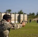 A person in military camouflage uniform is firing a semi-automatic pistol at an outdoor shooting range. The shooter is using a two-handed grip stance aimed at silhouette targets downrange. The setting appears to be a military or law enforcement training facility with a berm and target stands visible in the background.