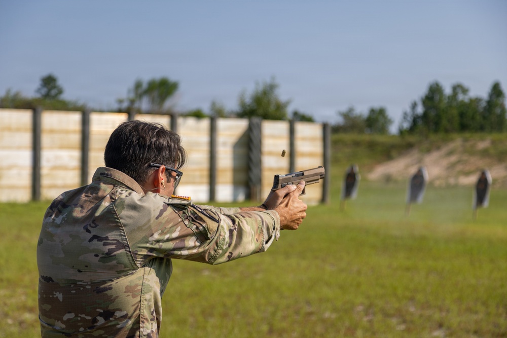 A person wearing military camouflage uniform and protective eyewear is firing a semi-automatic pistol at an outdoor shooting range. A spent brass casing is visibly ejecting from the firearm mid-shot. Multiple silhouette targets are positioned downrange against a wooden berm backdrop.