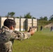 A person wearing military camouflage uniform and protective eyewear is firing a semi-automatic pistol at an outdoor shooting range. A spent brass casing is visibly ejecting from the firearm mid-shot. Multiple silhouette targets are positioned downrange against a wooden berm backdrop.