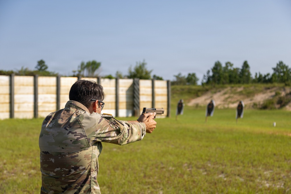A person wearing military camouflage uniform and protective eyewear is firing a semi-automatic pistol at a shooting range. Multiple silhouette targets are visible downrange against a berm backdrop. The setting appears to be an outdoor military or law enforcement training facility.