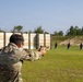 A person wearing military camouflage uniform and protective eyewear is firing a semi-automatic pistol at a shooting range. Multiple silhouette targets are visible downrange against a berm backdrop. The setting appears to be an outdoor military or law enforcement training facility.