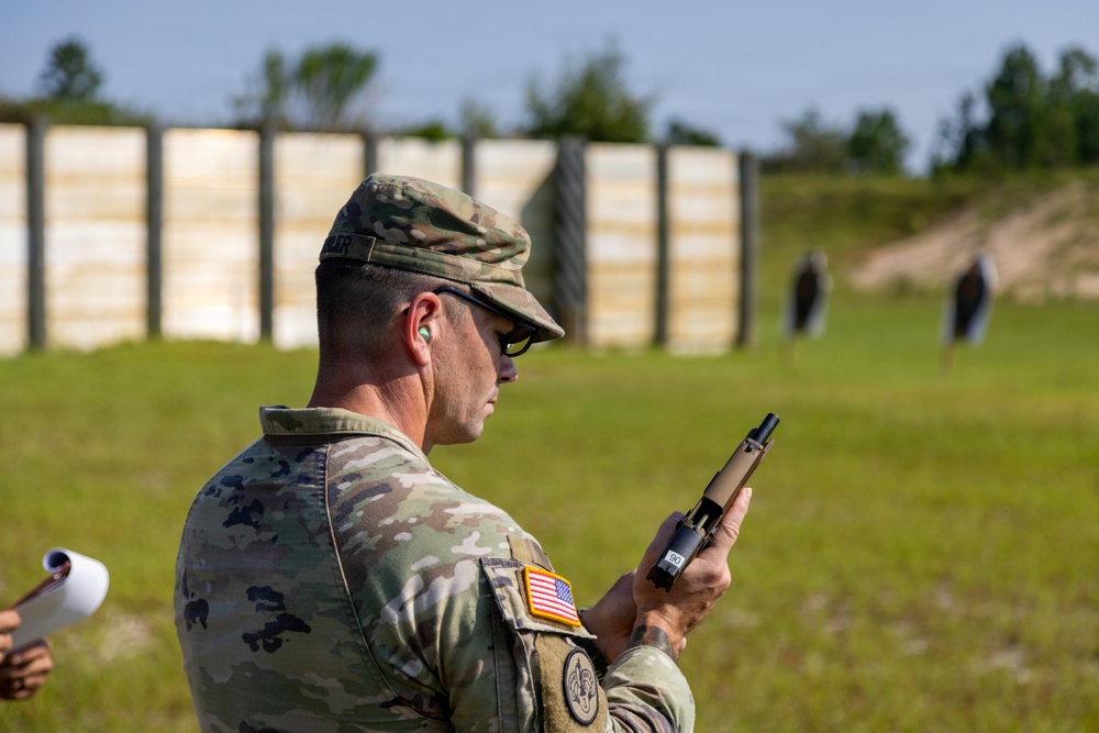 A U.S. Army soldier in MultiCam (OCP) uniform is standing at what appears to be a military shooting range, inspecting or handling a pistol. The soldier is wearing hearing protection (ear plugs) and sunglasses, with an American flag patch and what appears to be a unit patch visible on the left sleeve. Silhouette targets are visible downrange in the background.