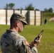 A U.S. Army soldier in MultiCam (OCP) uniform is standing at what appears to be a military shooting range, inspecting or handling a pistol. The soldier is wearing hearing protection (ear plugs) and sunglasses, with an American flag patch and what appears to be a unit patch visible on the left sleeve. Silhouette targets are visible downrange in the background.