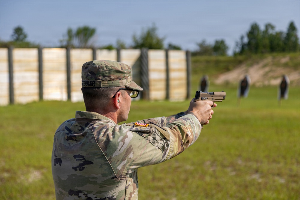 A U.S. Army soldier in Operational Camouflage Pattern (OCP) uniform is firing a pistol at a military shooting range. The soldier is in a two-handed shooting stance, aiming at multiple silhouette targets downrange. The setting appears to be an outdoor live-fire training range with wooden barriers and a berm in the background.