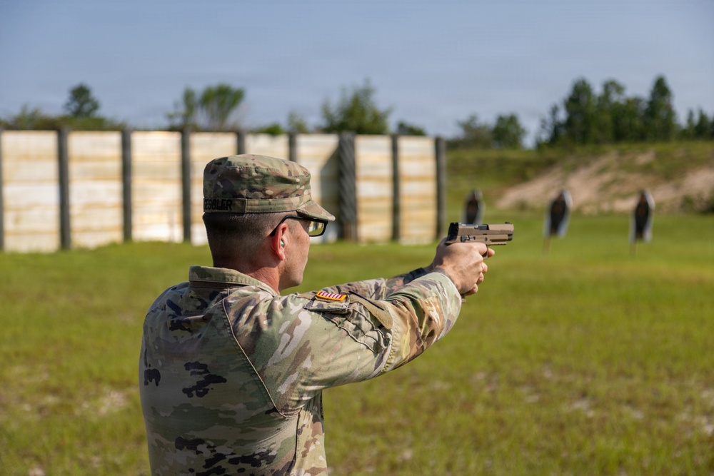 A U.S. Army soldier in Operational Camouflage Pattern (OCP) uniform is firing a pistol at a military shooting range. The soldier is in a two-handed shooting stance, aiming downrange at multiple silhouette targets. The setting appears to be an outdoor qualification or training range with a wooden backstop barrier visible in the background.
