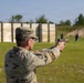 A U.S. Army soldier in Operational Camouflage Pattern (OCP) uniform is firing a pistol at a military shooting range. The soldier is in a two-handed shooting stance, aiming downrange at multiple silhouette targets. The setting appears to be an outdoor qualification or training range with a wooden backstop barrier visible in the background.