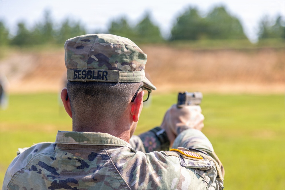 A U.S. Army soldier named Gessler is photographed from behind at what appears to be an outdoor firing range. The soldier is in a shooting stance, aiming a pistol downrange while wearing OCP (Operational Camouflage Pattern) uniform. A U.S. flag patch and what appears to be a unit patch are visible on the right sleeve.
