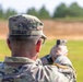A U.S. Army soldier named Gessler is photographed from behind at what appears to be an outdoor firing range. The soldier is in a shooting stance, aiming a pistol downrange while wearing OCP (Operational Camouflage Pattern) uniform. A U.S. flag patch and what appears to be a unit patch are visible on the right sleeve.