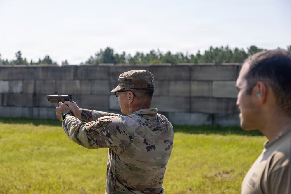 A soldier in U.S. Army OCP (Operational Camouflage Pattern) uniform is firing a pistol at an outdoor shooting range. A second individual in a tan/olive shirt stands nearby observing, likely acting as a range safety officer or instructor. Both individuals are wearing ear protection, consistent with standard range safety protocols.