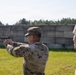 A soldier in U.S. Army OCP (Operational Camouflage Pattern) uniform is firing a pistol at an outdoor shooting range. A second individual in a tan/olive shirt stands nearby observing, likely acting as a range safety officer or instructor. Both individuals are wearing ear protection, consistent with standard range safety protocols.