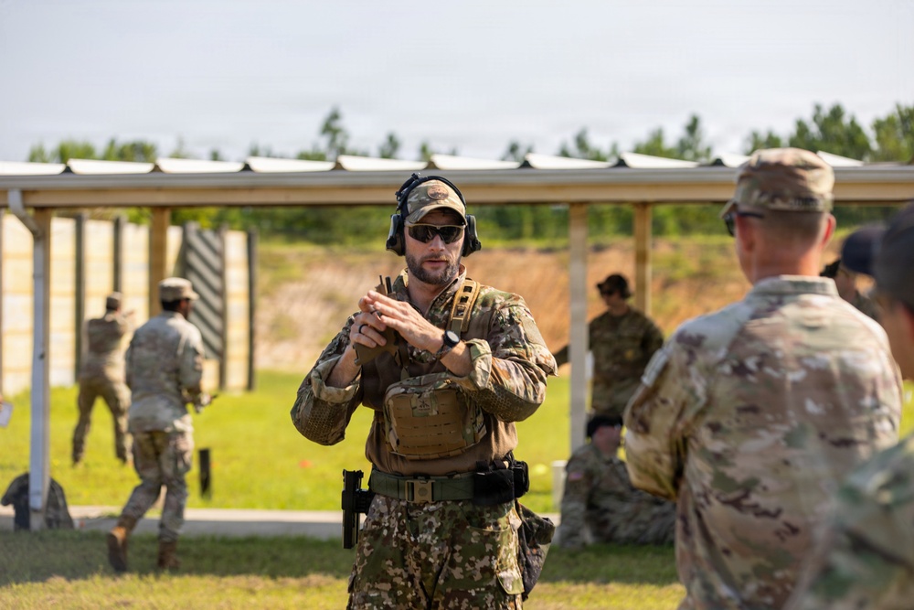 A military firearms training session is taking place at an outdoor shooting range. A central instructor in full tactical gear, including a plate carrier and hearing protection, appears to be demonstrating a weapon handling technique to other personnel. Multiple soldiers in camouflage uniforms are visible in the background actively engaging targets on the range.