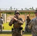 A military firearms training session is taking place at an outdoor shooting range. A central instructor in full tactical gear, including a plate carrier and hearing protection, appears to be demonstrating a weapon handling technique to other personnel. Multiple soldiers in camouflage uniforms are visible in the background actively engaging targets on the range.