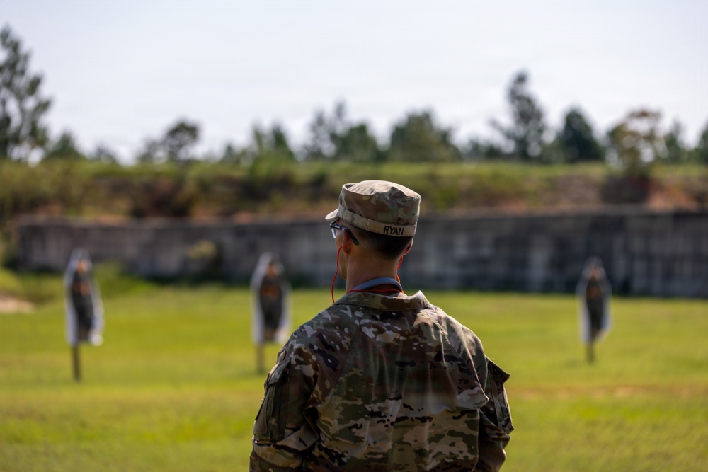 A soldier wearing MultiCam/OCP camouflage uniform with the name tape 'RYAN' is observed from behind, standing on a firing range. The soldier is wearing eye protection and hearing protection (red cord visible), facing multiple human-silhouette targets downrange. The setting appears to be an outdoor military shooting range with a berm backstop and treeline in the background.