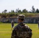 A soldier wearing MultiCam/OCP camouflage uniform with the name tape 'RYAN' is observed from behind, standing on a firing range. The soldier is wearing eye protection and hearing protection (red cord visible), facing multiple human-silhouette targets downrange. The setting appears to be an outdoor military shooting range with a berm backstop and treeline in the background.