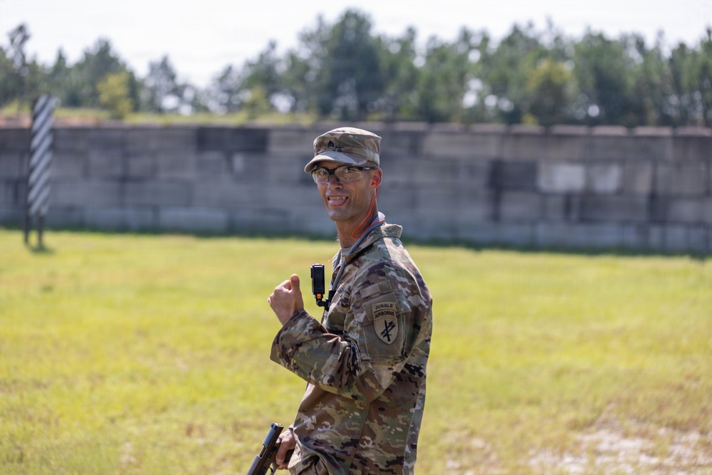 A U.S. Army soldier in MultiCam (OCP) camouflage uniform is standing outdoors on what appears to be a military training range or installation. The soldier is wearing a 'Jungle Airborne' patch on the right shoulder and is holding what appears to be a firearm at their side while giving a thumbs-up gesture. The background shows a concrete berm or wall typical of a shooting range, with trees visible beyond it.