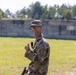 A U.S. Army soldier in MultiCam (OCP) camouflage uniform is standing outdoors on what appears to be a military training range or installation. The soldier is wearing a 'Jungle Airborne' patch on the right shoulder and is holding what appears to be a firearm at their side while giving a thumbs-up gesture. The background shows a concrete berm or wall typical of a shooting range, with trees visible beyond it.