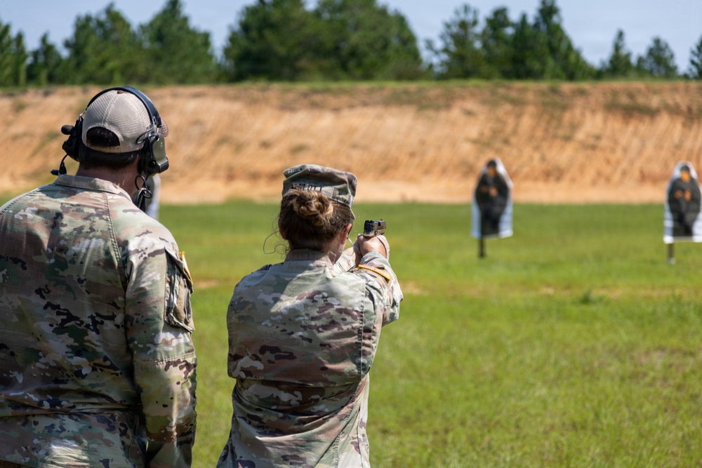 Two military personnel in MultiCam/OCP camouflage uniforms are at an outdoor shooting range. One soldier is actively firing a pistol at humanoid silhouette targets downrange, while the other observes wearing hearing protection. The setting appears to be a standard military qualification or training range with a dirt berm backstop and tree line in the background.