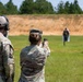 Two military personnel in MultiCam/OCP camouflage uniforms are at an outdoor shooting range. One soldier is actively firing a pistol at humanoid silhouette targets downrange, while the other observes wearing hearing protection. The setting appears to be a standard military qualification or training range with a dirt berm backstop and tree line in the background.